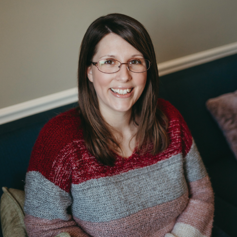 A woman with brown hair wearing glasses and a striped sweater, sitting on a couch and smiling.