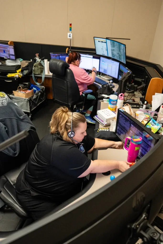 Two female operators working at multiple computer monitors in a control room, surrounded by various items and supplies.