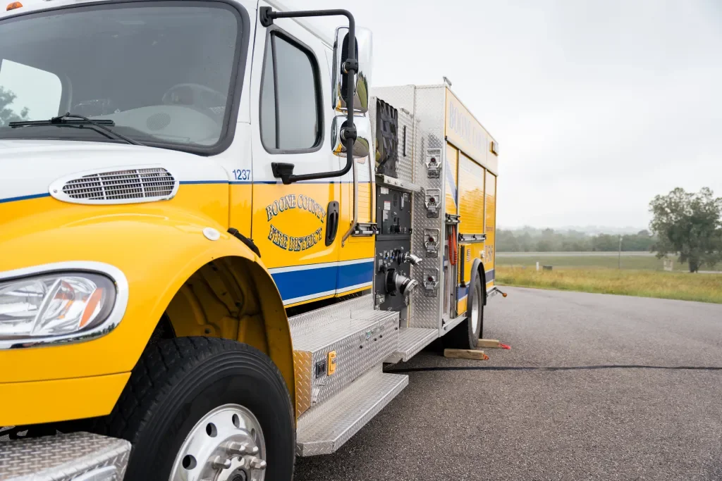 Yellow and white emergency fire truck with blue stripe, parked on the side of a paved road, with trees and grass in the background.