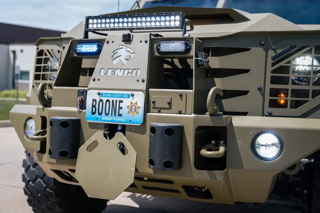 Close-up of a tan armored police vehicle with a Missouri license plate from Boone County Sheriff's Office, featuring a logo of a panther and mounted lights, grille, and bumper.