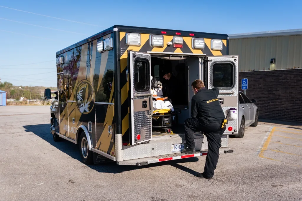 Paramedics preparing a patient on a stretcher in an ambulance in a parking lot outside a building.