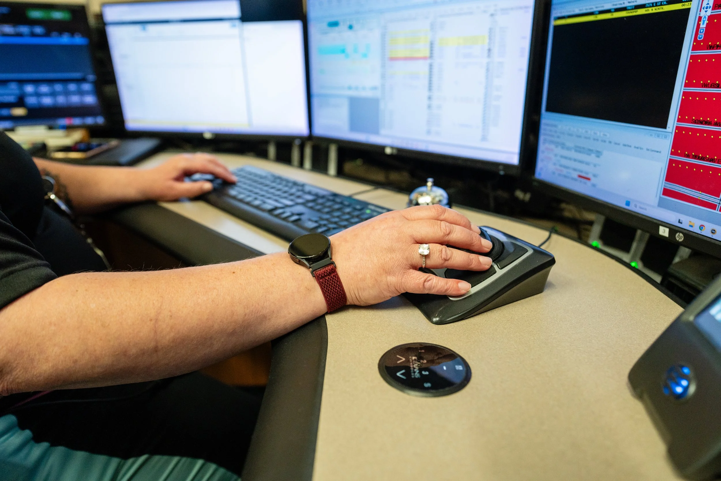 Person using a computer mouse and keyboard in front of multiple monitors displaying data and graphs, possibly in a control room or office setting.
