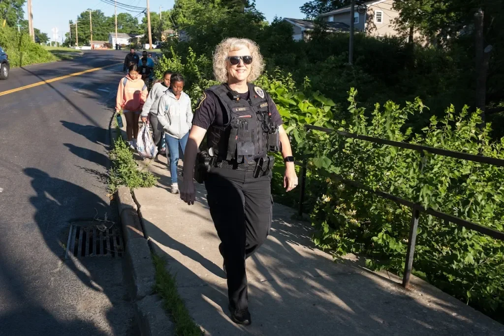 A female police officer with sunglasses leading a group of people, including children, on a walk on a sidewalk in a suburban neighborhood on a sunny day.