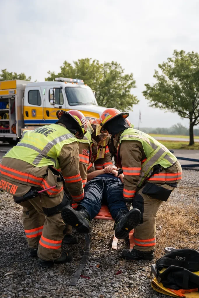 Four firefighters in yellow and orange gear carrying a person on a stretcher with a fire truck in the background.