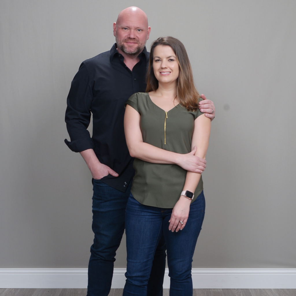 A man and woman standing together in front of a plain gray wall, smiling at the camera.