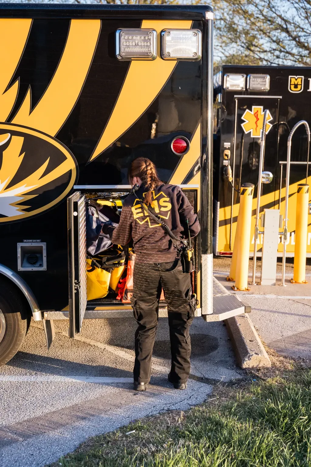 Paramedic with braided hair, dressed in a black EMS uniform, standing outside a black and yellow EMS vehicle, reaching into an open storage compartment.