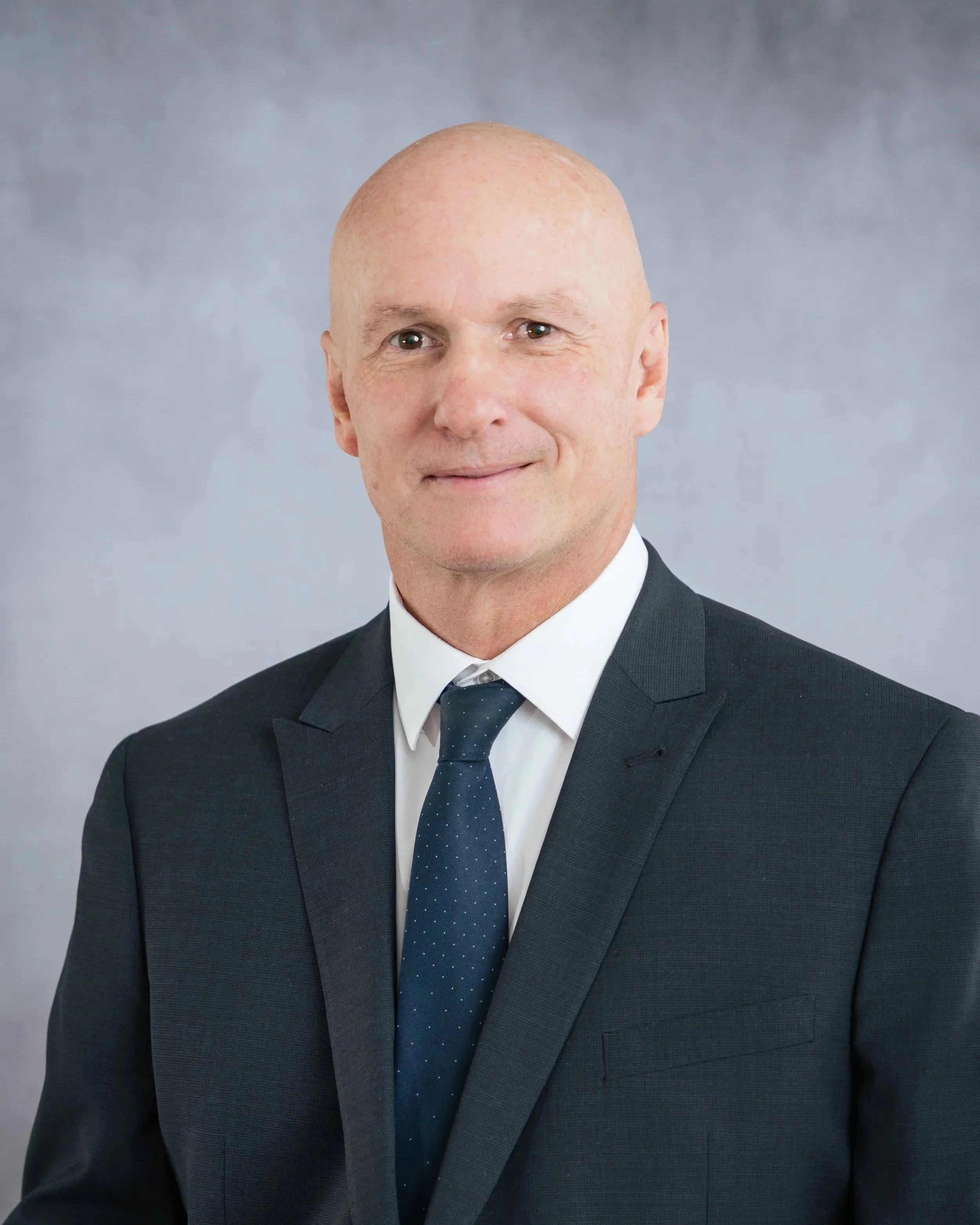 Headshot of a bald man in a black suit, white shirt, and navy blue tie with white dots, smiling subtly against a gray background.