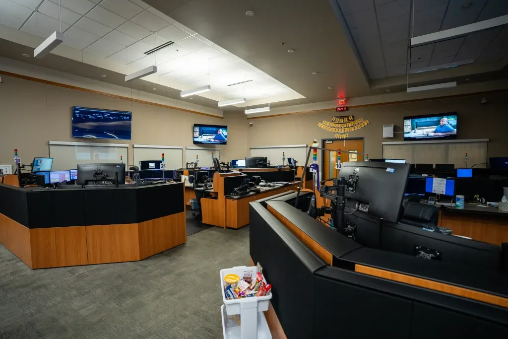A call center with multiple workstations, monitors, and televisions on the walls displaying a news program, with some desks equipped with phones and headsets.
