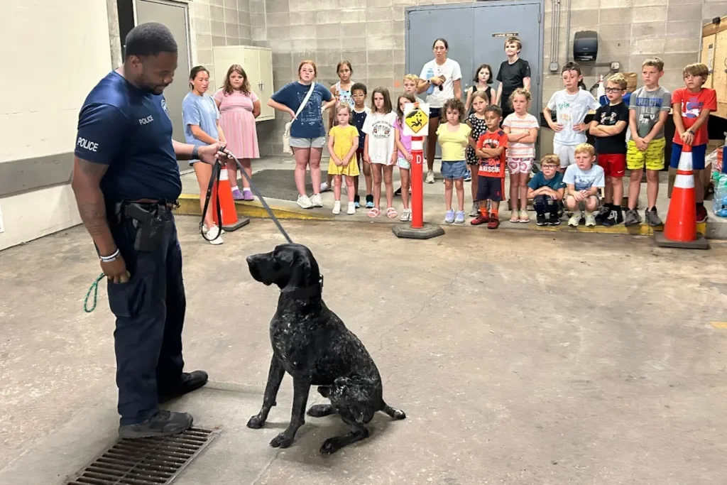 A police officer with a black police dog in an indoor area with children watching in the background.