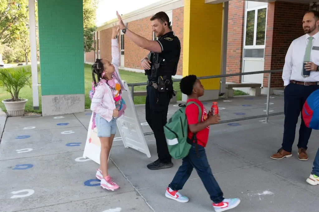 A police officer high-fiving a young girl while two boys walk by, outside a school building.