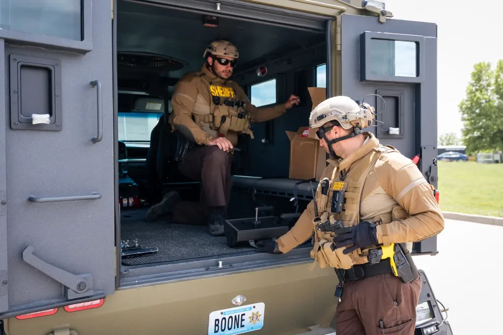 Two law enforcement officers in tactical gear, one inside a military vehicle and the other outside, preparing for a situation. The vehicle has a license plate that says Boone.