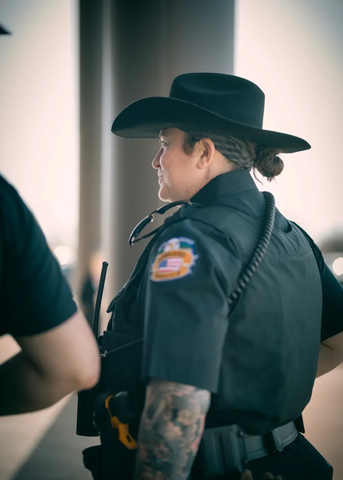 Profile of a police officer in uniform with a patch, wearing a cowboy hat, engaged in conversation indoors.