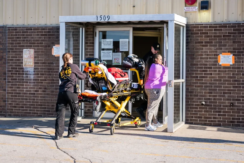A person in a black jacket and a woman in a purple jacket enter a building with a green medical stretcher parked outside. An emergency responder is holding the stretcher, and there are signs on the building's wall.
