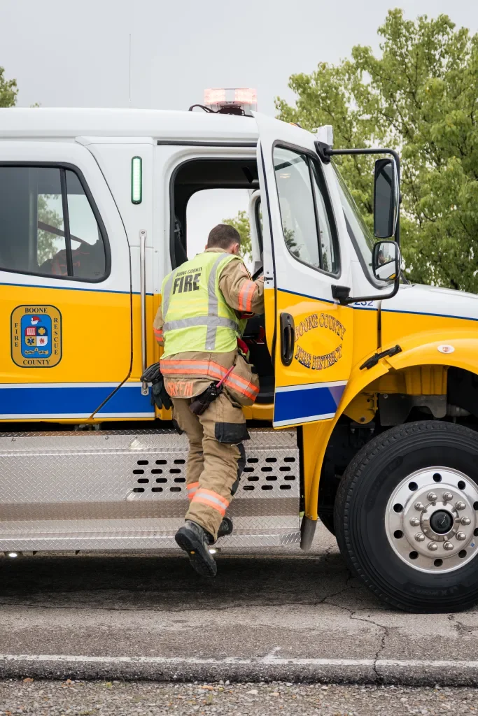 A firefighter in uniform and reflective vest climbing into a Boone County Fire Rescue vehicle, which is a large yellow and white emergency truck.