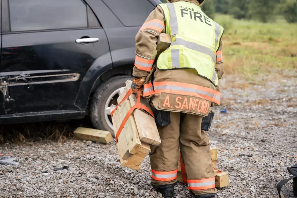 Firefighter wearing a vest with 'FIRE' and 'A. SANFORD' on it, carrying wooden blocks near a damaged black vehicle on gravel ground.