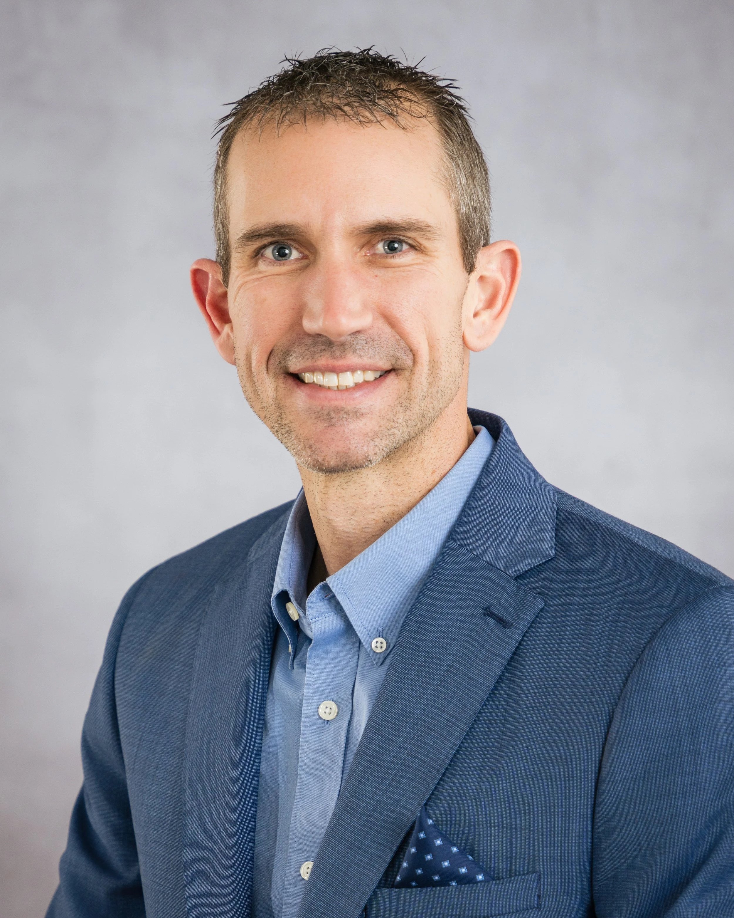 Headshot of a man in a blue suit with a light blue shirt, smiling, against a plain gray background.