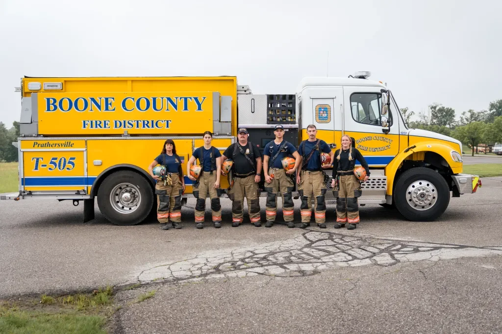Group of six firefighters standing in front of a Boone County Fire District truck, holding helmets and dressed in firefighting gear.