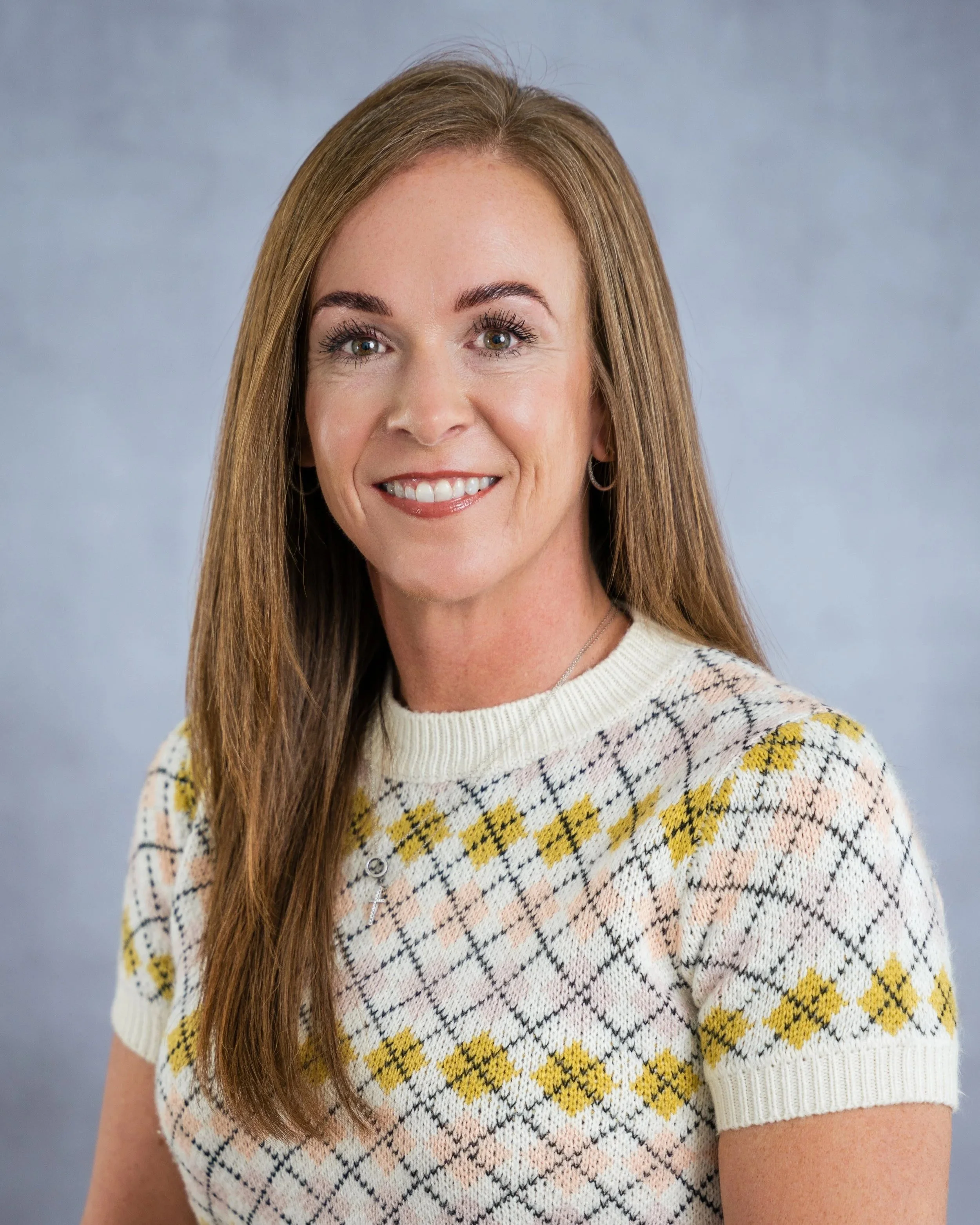 A woman with long brown hair and light skin, smiling, wearing a cream sweater with a yellow and black argyle pattern, against a gradient gray background.
