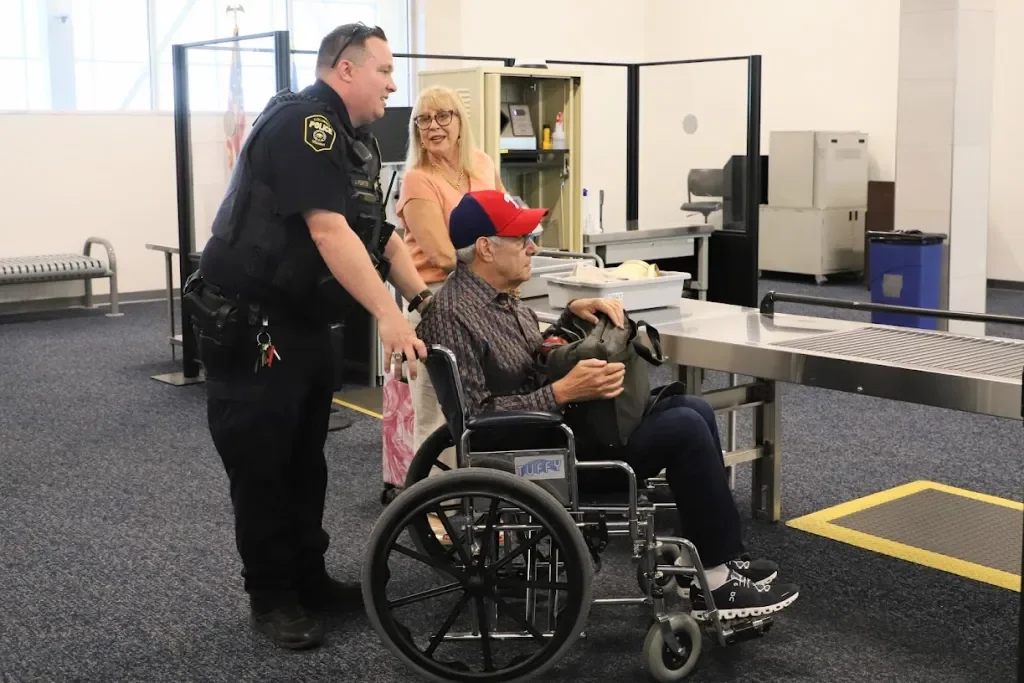 A woman and a man in a wheelchair at an airport security checkpoint, with a security officer talking to the man. The woman is smiling and looking at the officer. The man is wearing a red cap and sunglasses, holding a backpack.