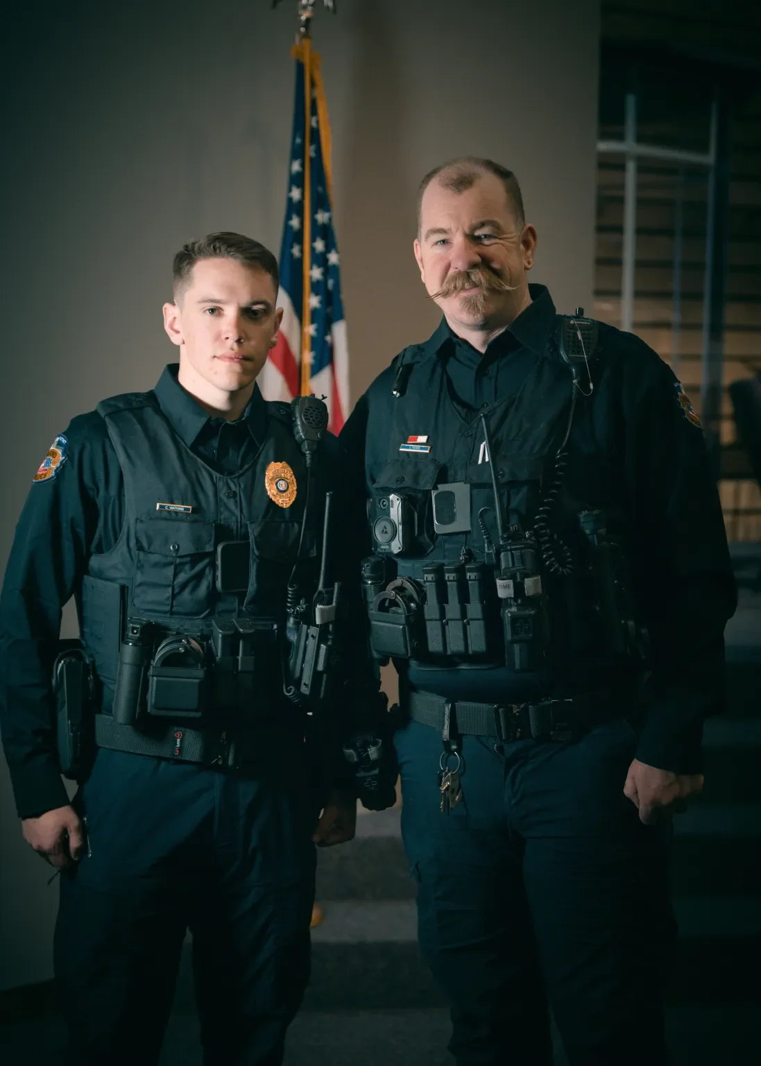 Two police officers in uniform standing together inside a building, with an American flag in the background.