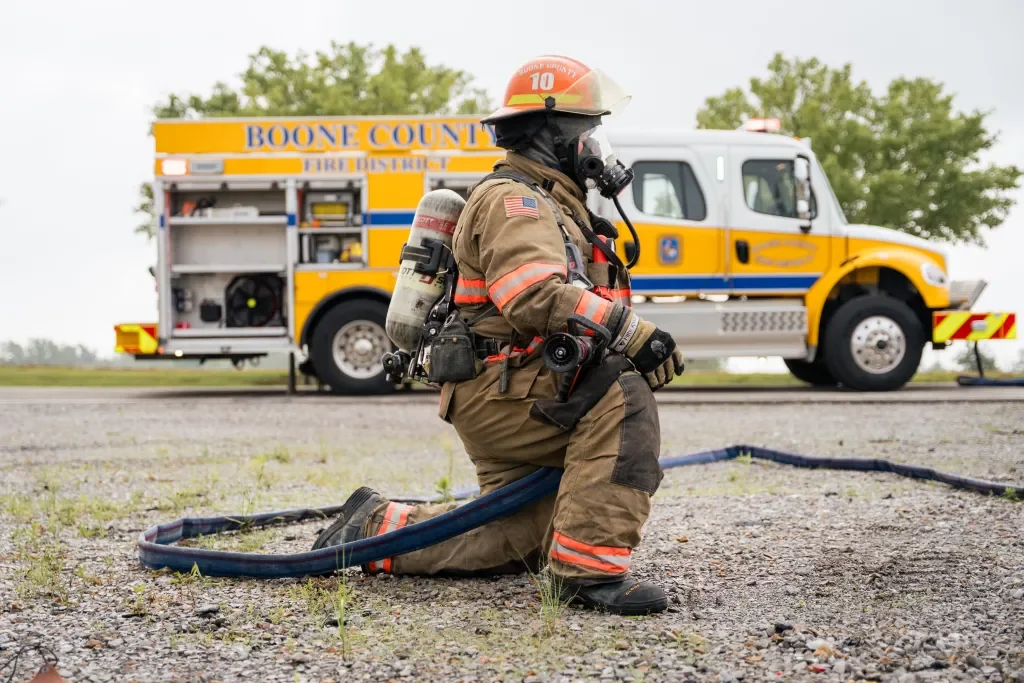 A firefighter kneeling on the ground, dressed in full gear including an oxygen mask, with a Boone County fire truck and a yellow emergency vehicle in the background.