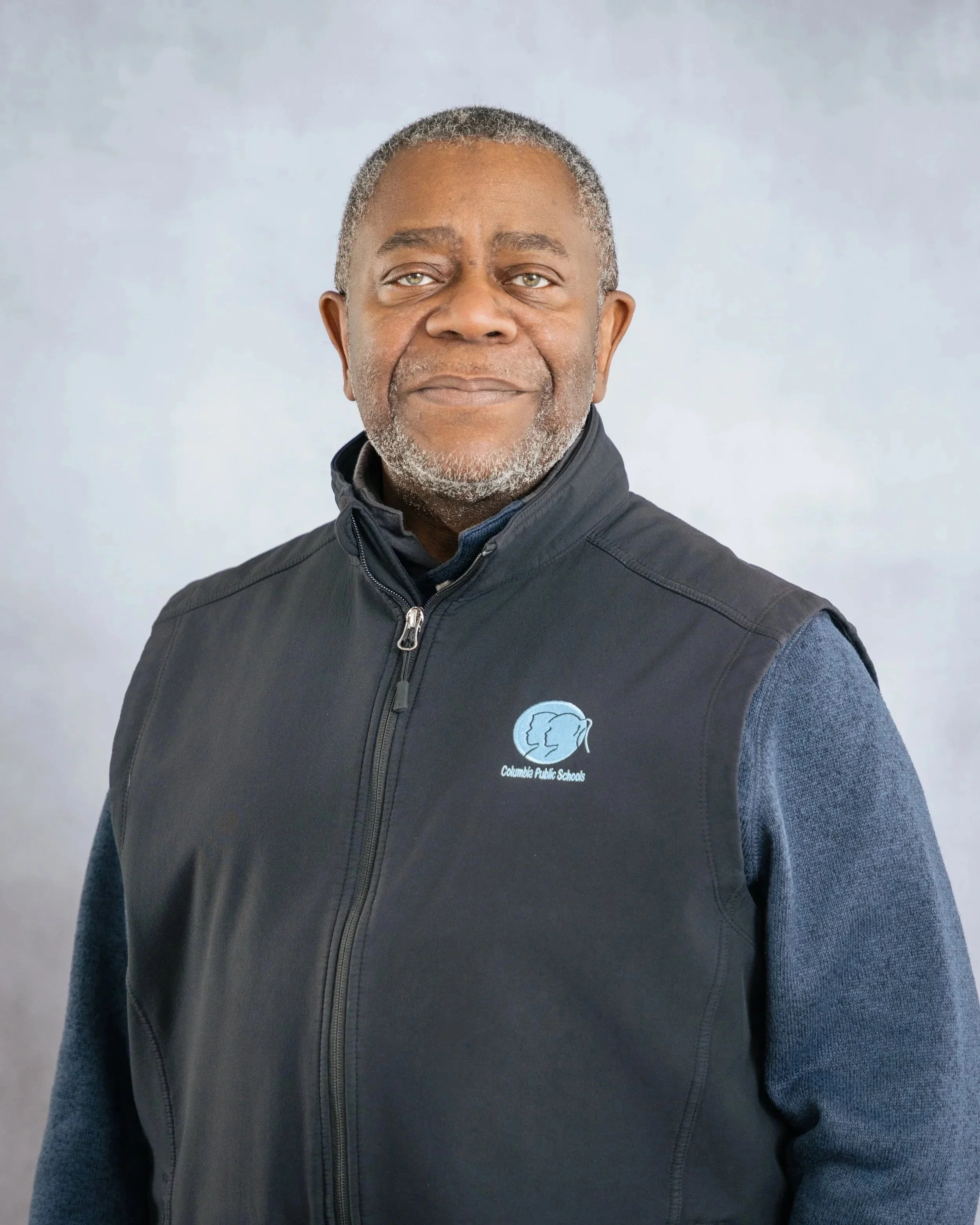 Portrait of a senior man with gray hair and beard, wearing a Columbia Public Schools vest, smiling lightly against a gray background.