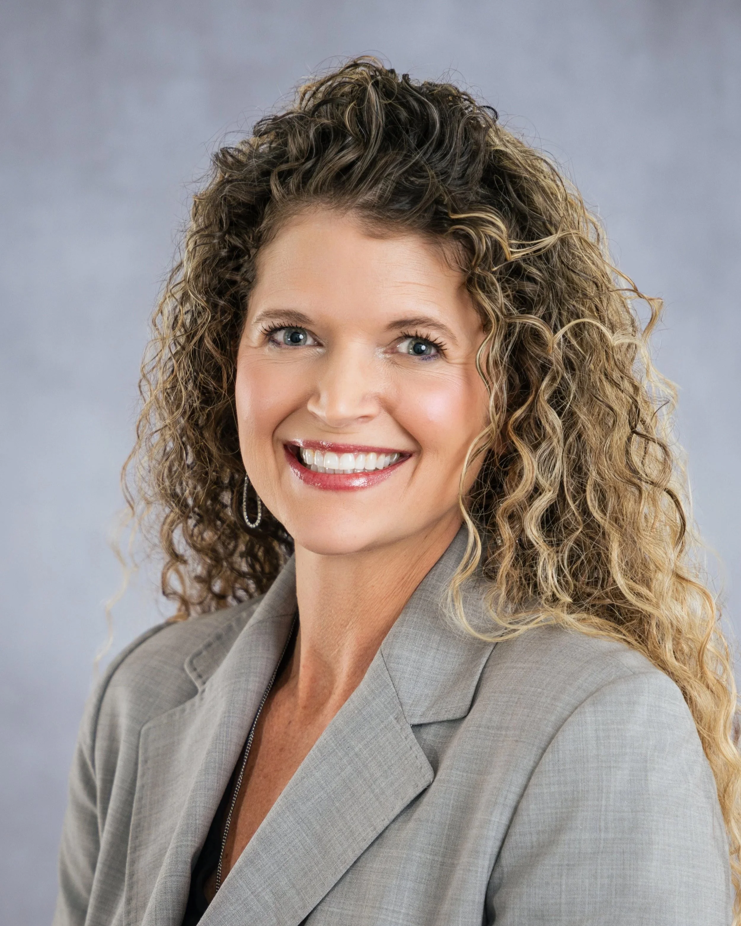 A woman with curly blonde hair wearing a gray blazer, smiling against a plain gray background.