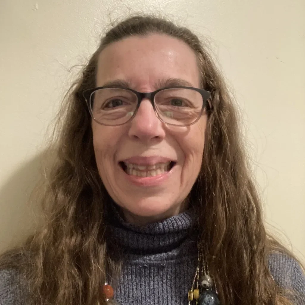 A woman with long, curly brown hair wearing glasses and a gray turtleneck sweater, smiling in front of a light-colored wall.
