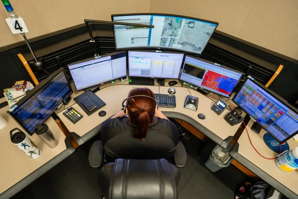 A person working at a multi-monitor control room station with multiple screens displaying maps, spreadsheets, and data.