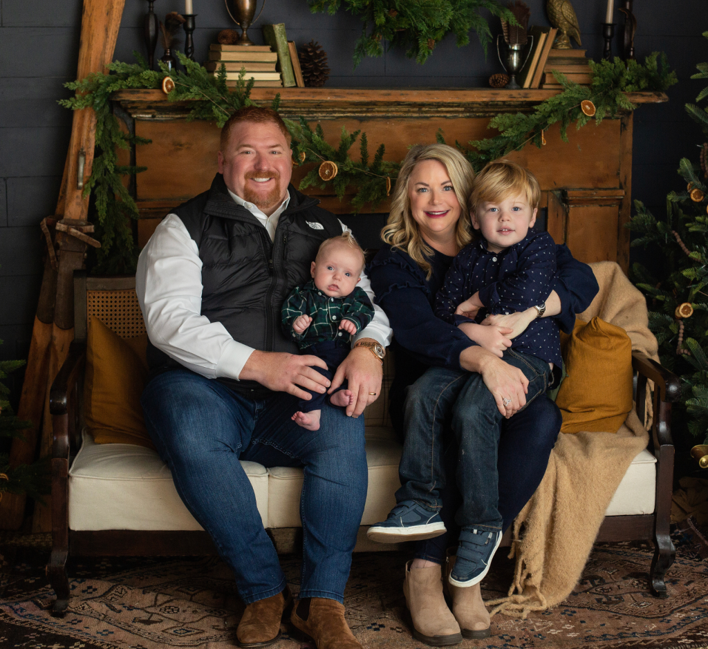 Family portrait of two adults and two children sitting on a vintage white and wood sofa with Christmas decorations in the background.