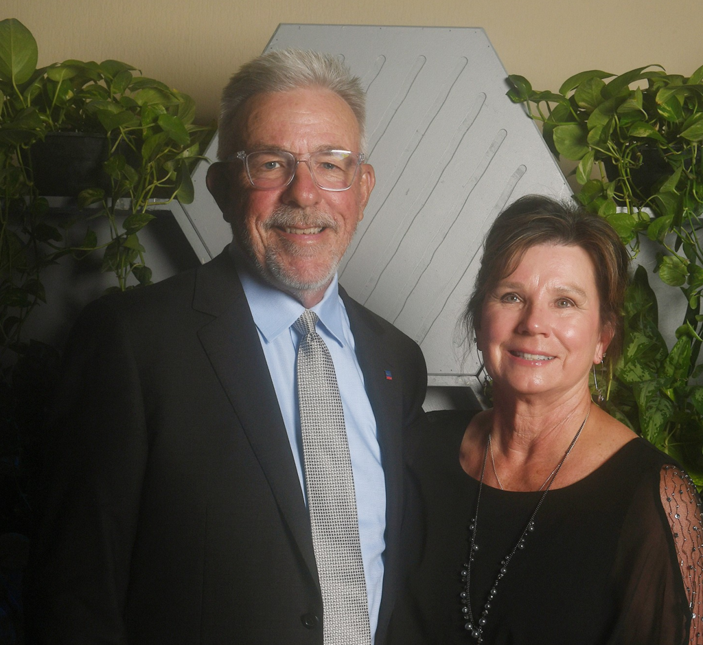 A middle-aged man and woman smiling and posing together indoors, with green plants and a hexagonal decor piece in the background.