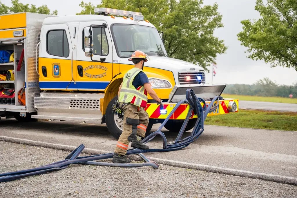 Firefighter in uniform and safety vest handling hoses near an emergency response truck with lights on, on a roadside with green trees in the background.