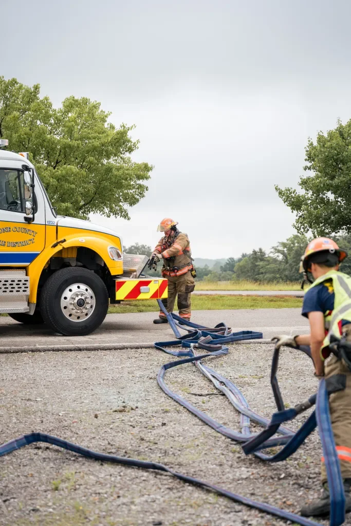 Firefighters connect hoses to a yellow fire truck at an outdoor scene with green trees and cloudy sky.