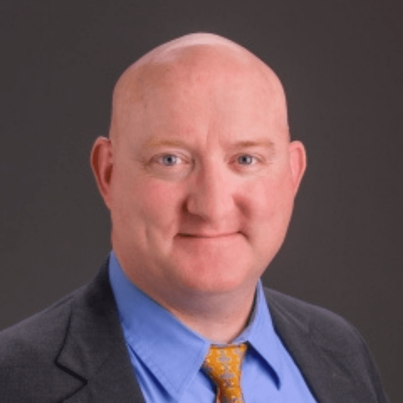 Professional headshot of a bald man wearing a gray suit, blue dress shirt, and yellow tie, smiling against a dark gray background.