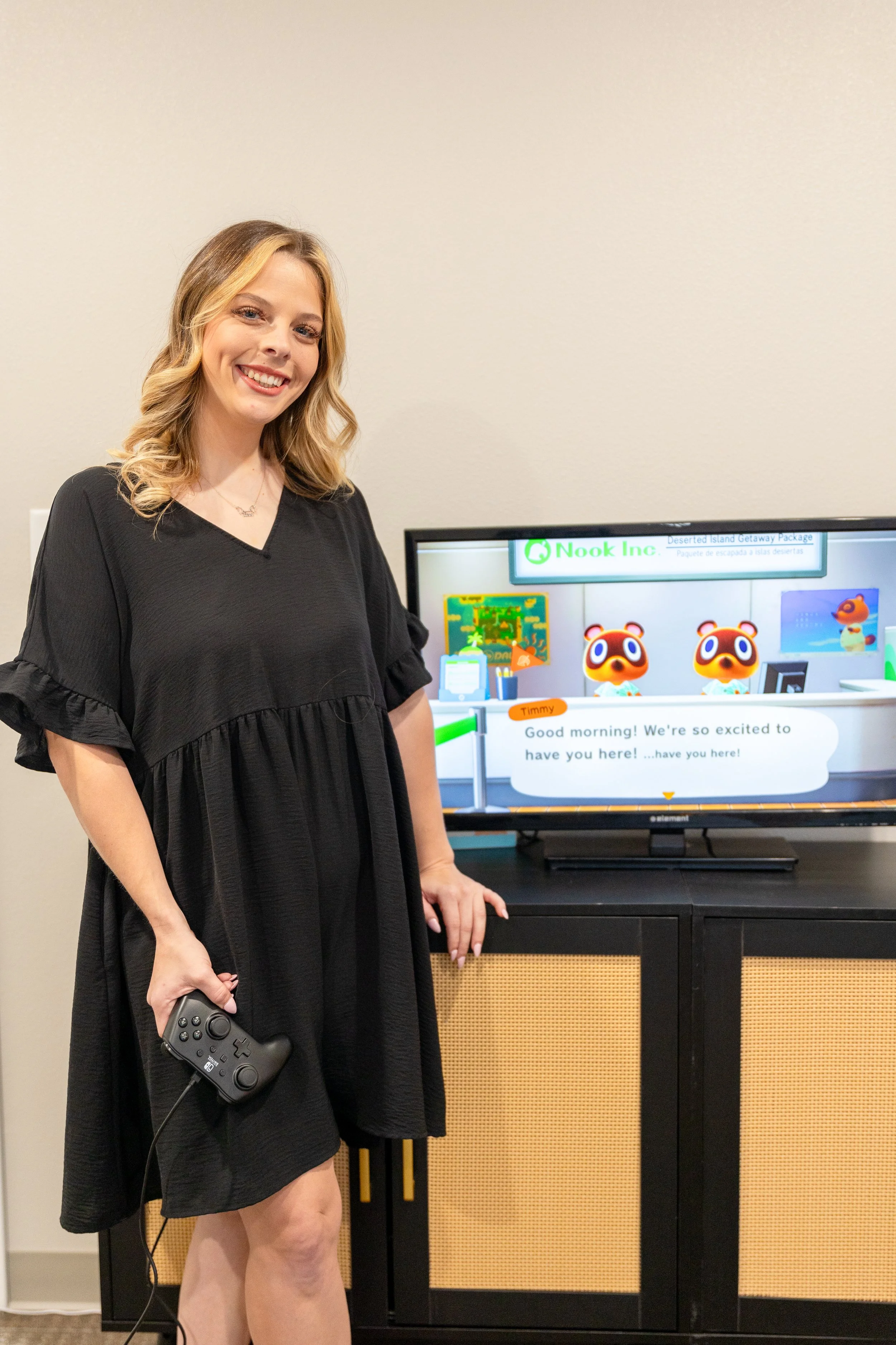 A woman with blonde, wavy hair wearing a black dress, holding a gaming controller, standing next to a TV monitor showing a scene from a video game featuring animated animals and a character, with a speech bubble.
