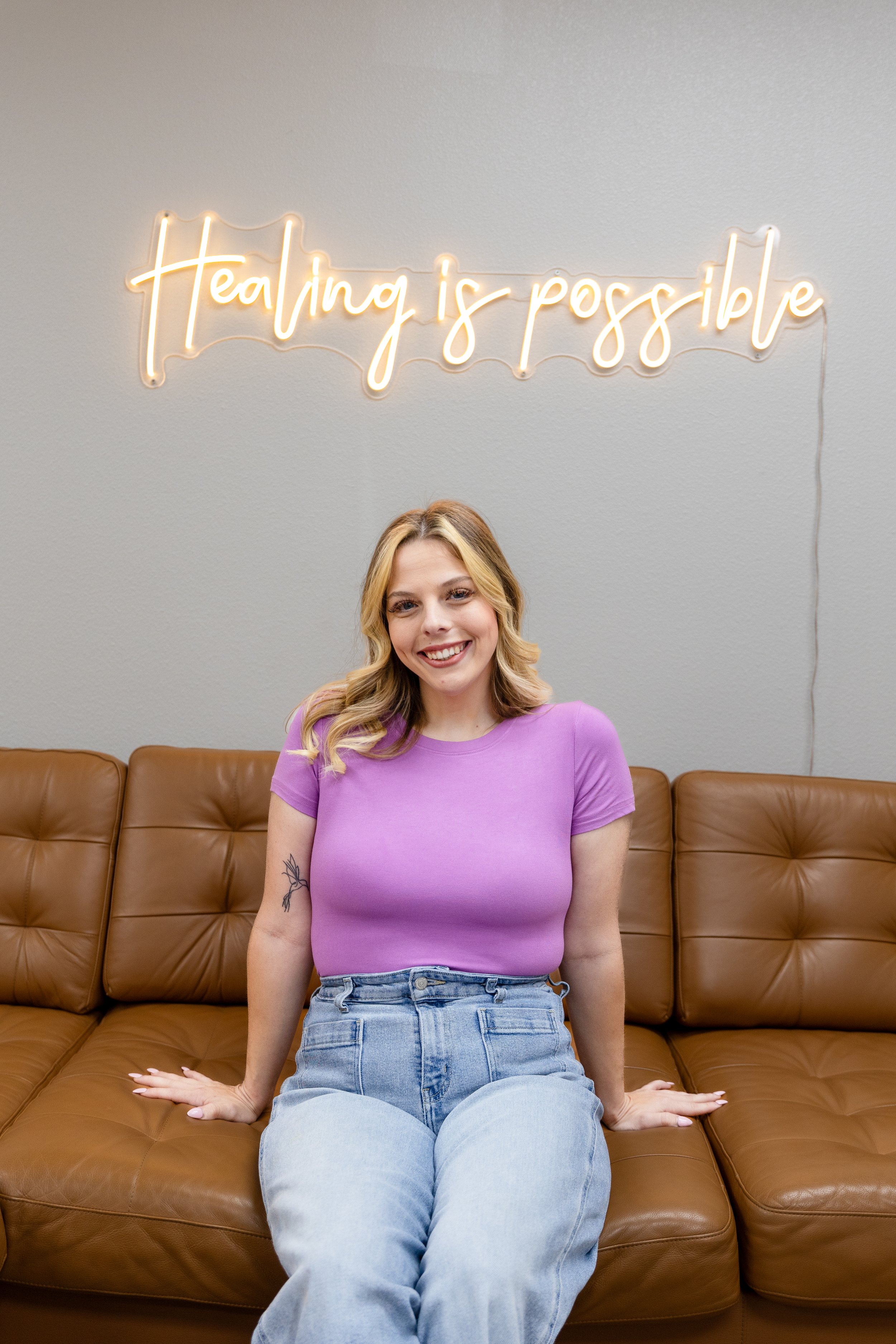 A young woman with blonde hair and a bright smile, wearing a purple top and light blue jeans, sitting on a brown leather couch. Behind her is a gray wall with a neon sign that reads 'Healing is possible'.
