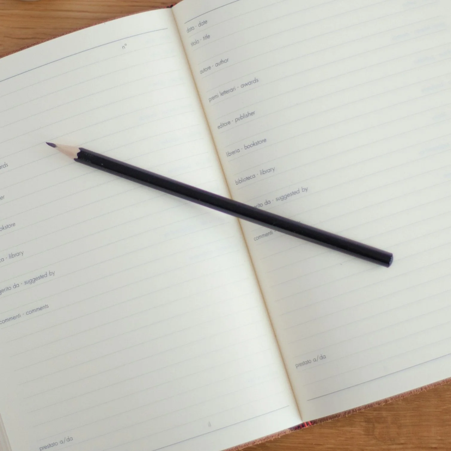 Open blank notebook with lined pages and a black pencil resting diagonally across the pages, placed on a wooden surface.