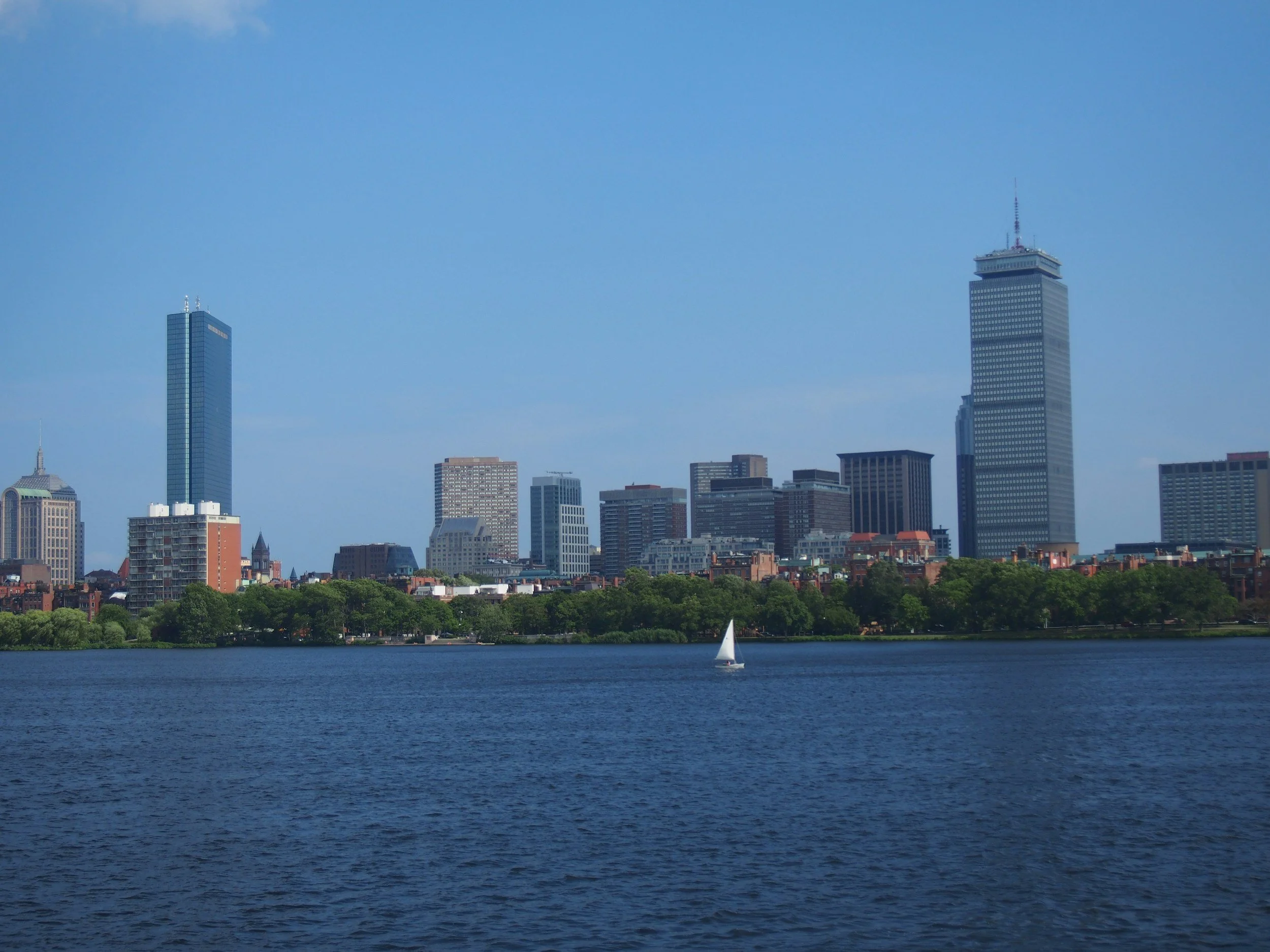 View of Boston skyline with tall skyscrapers, a body of water in the foreground, and a small sailboat on the water.