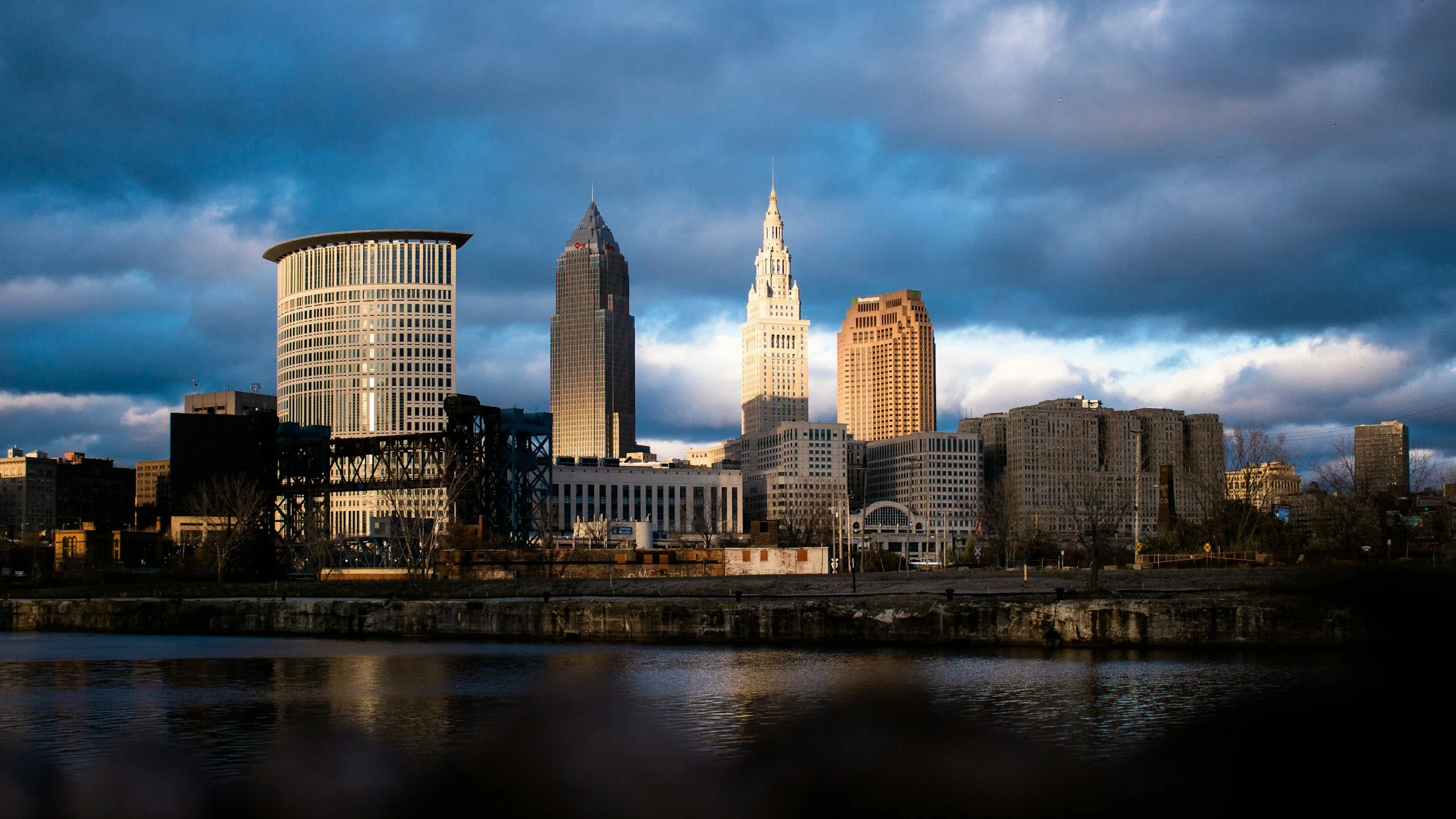 Skyline of Cleveland, Ohio with skyscrapers reflecting the late afternoon sunlight and a body of water in the foreground under cloudy skies.