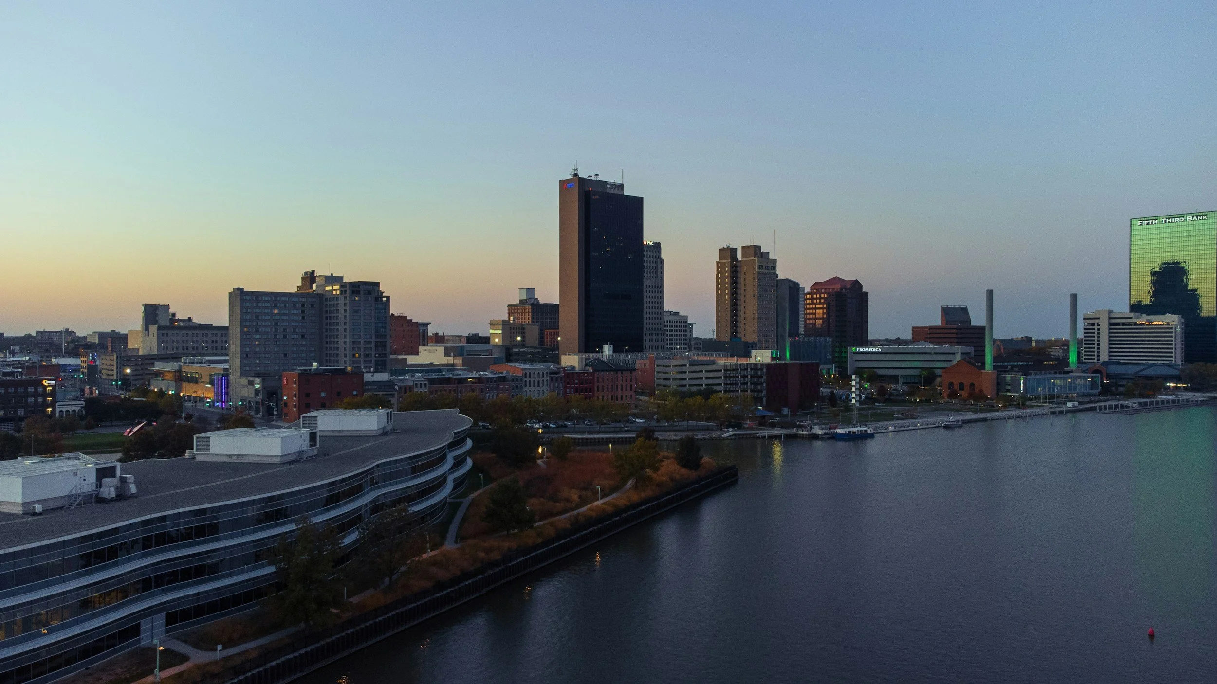 City skyline at dusk with buildings, water in the foreground, and a clear sky.