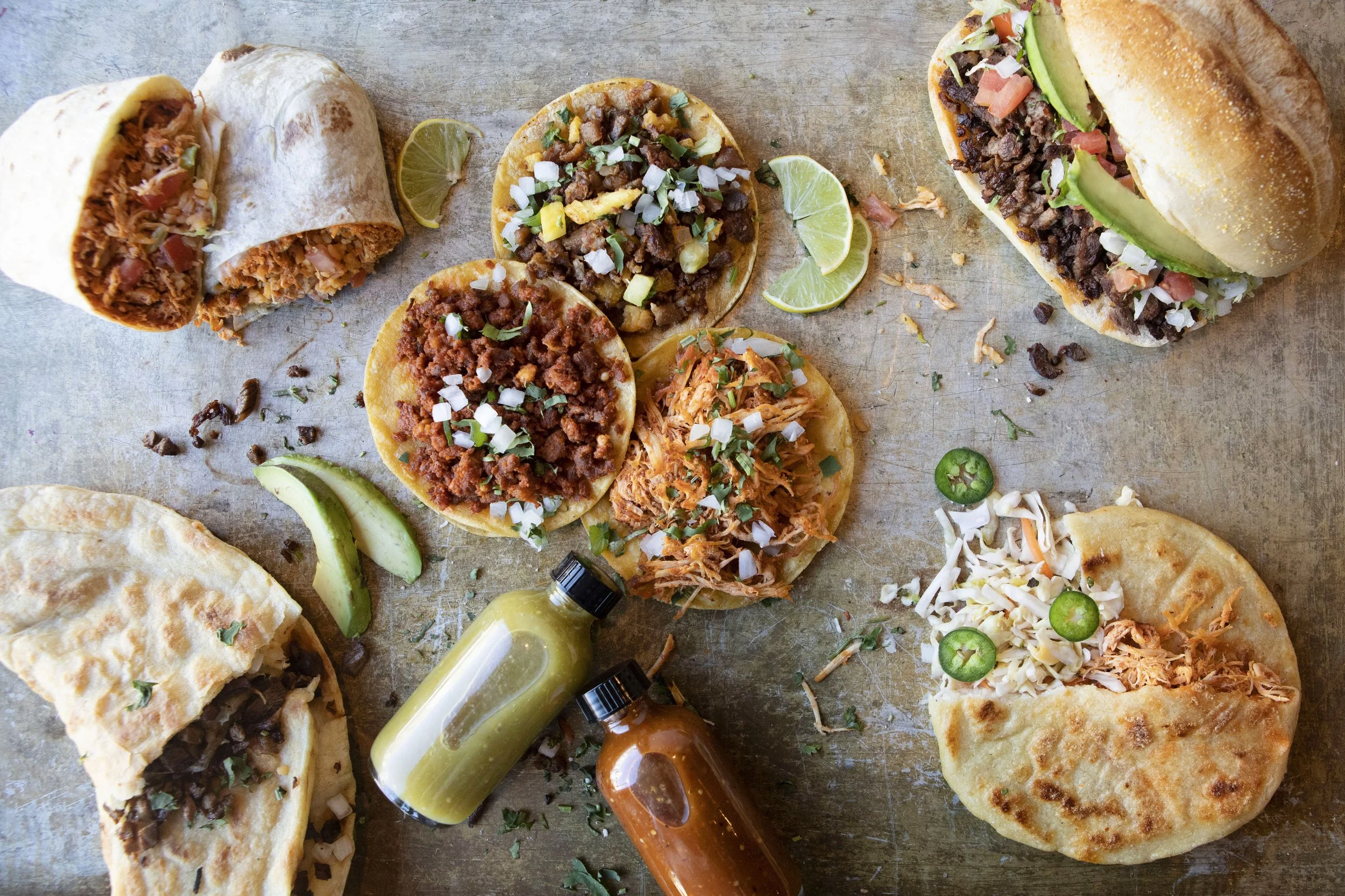 A variety of Mexican tacos with different fillings, sliced lime, and small bottles of hot sauce on a rustic surface.