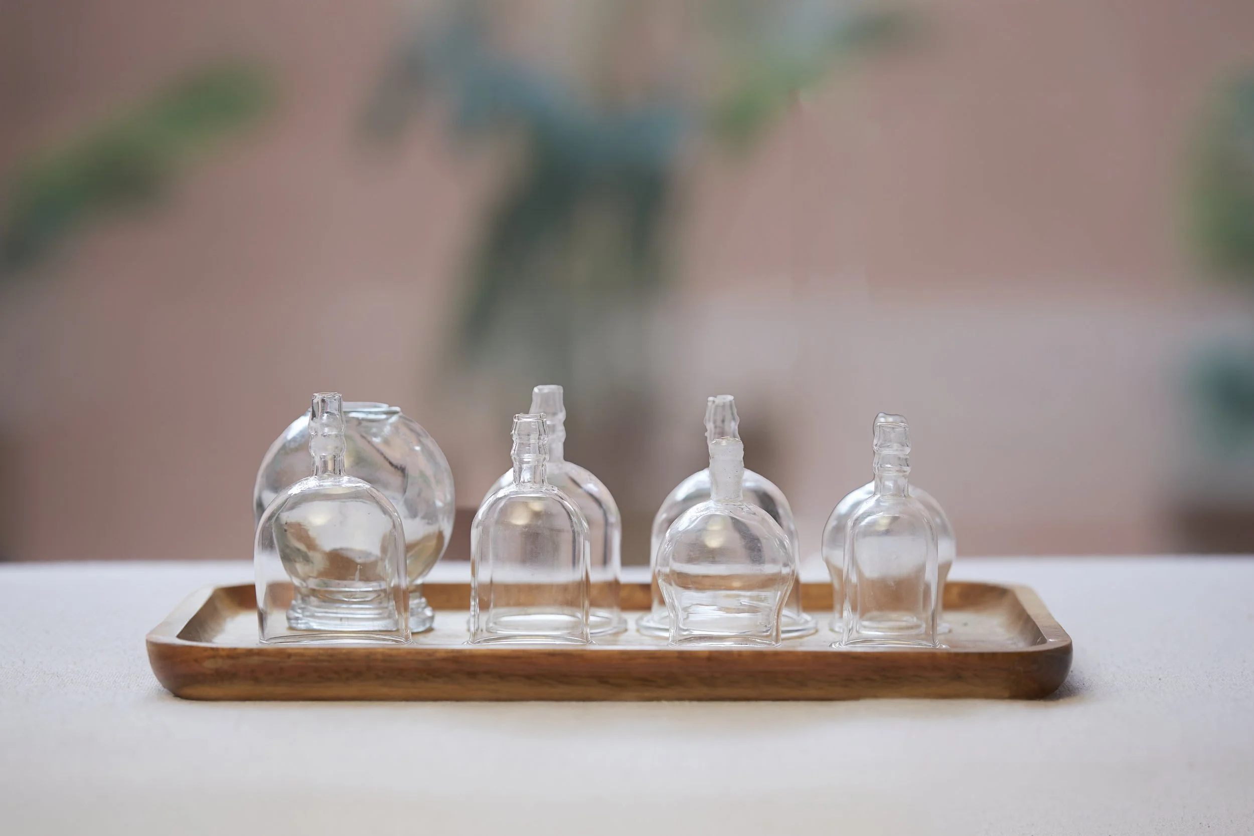 Six small empty glass bottles with varying shapes on a wooden tray, set on a white surface with a blurred background.