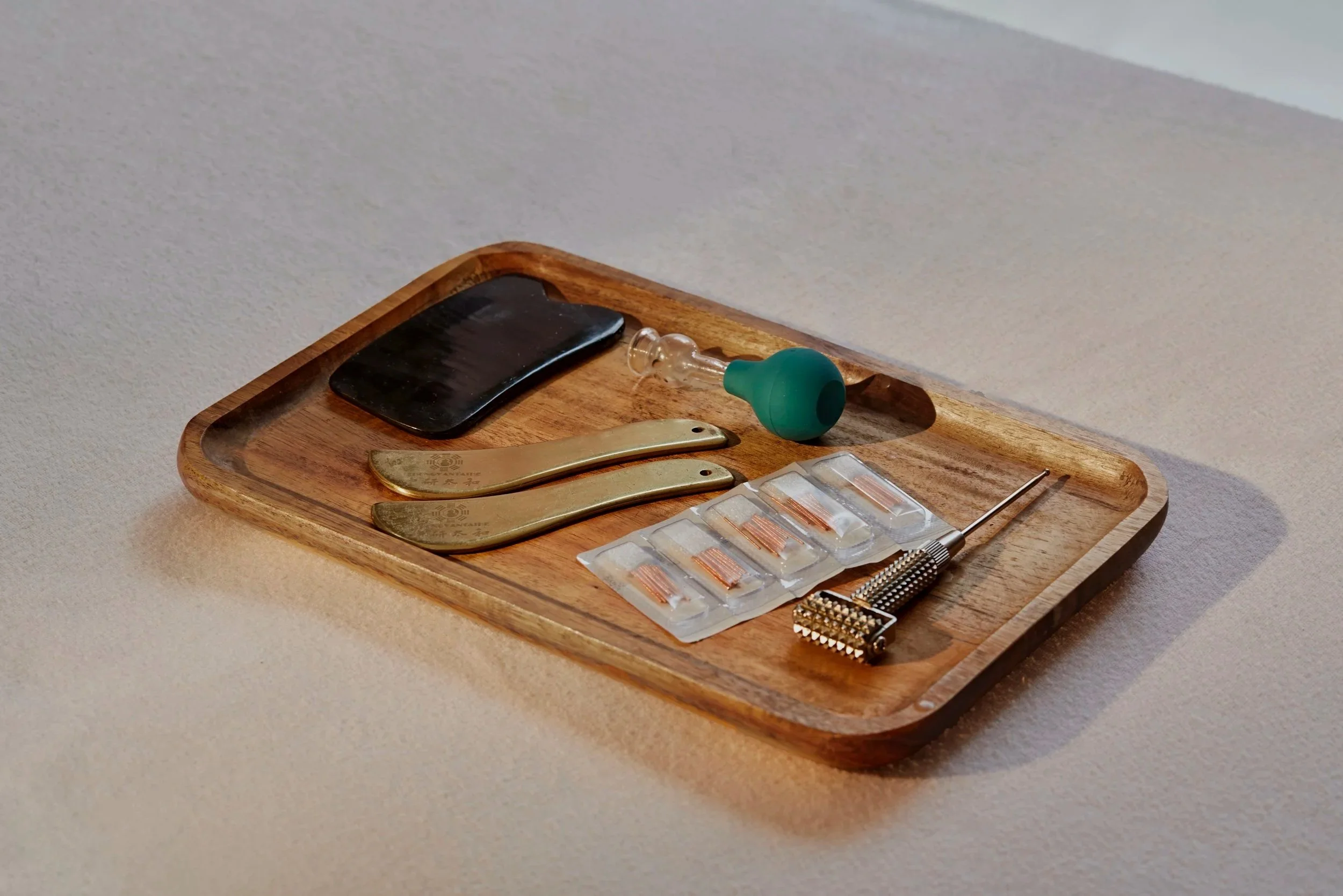 A wooden tray holding a black foot file, a green rubber bulb suction device, two gold-colored tools with engraved markings, a blister pack with small copper wire coils, and a metal tool with a textured handle.