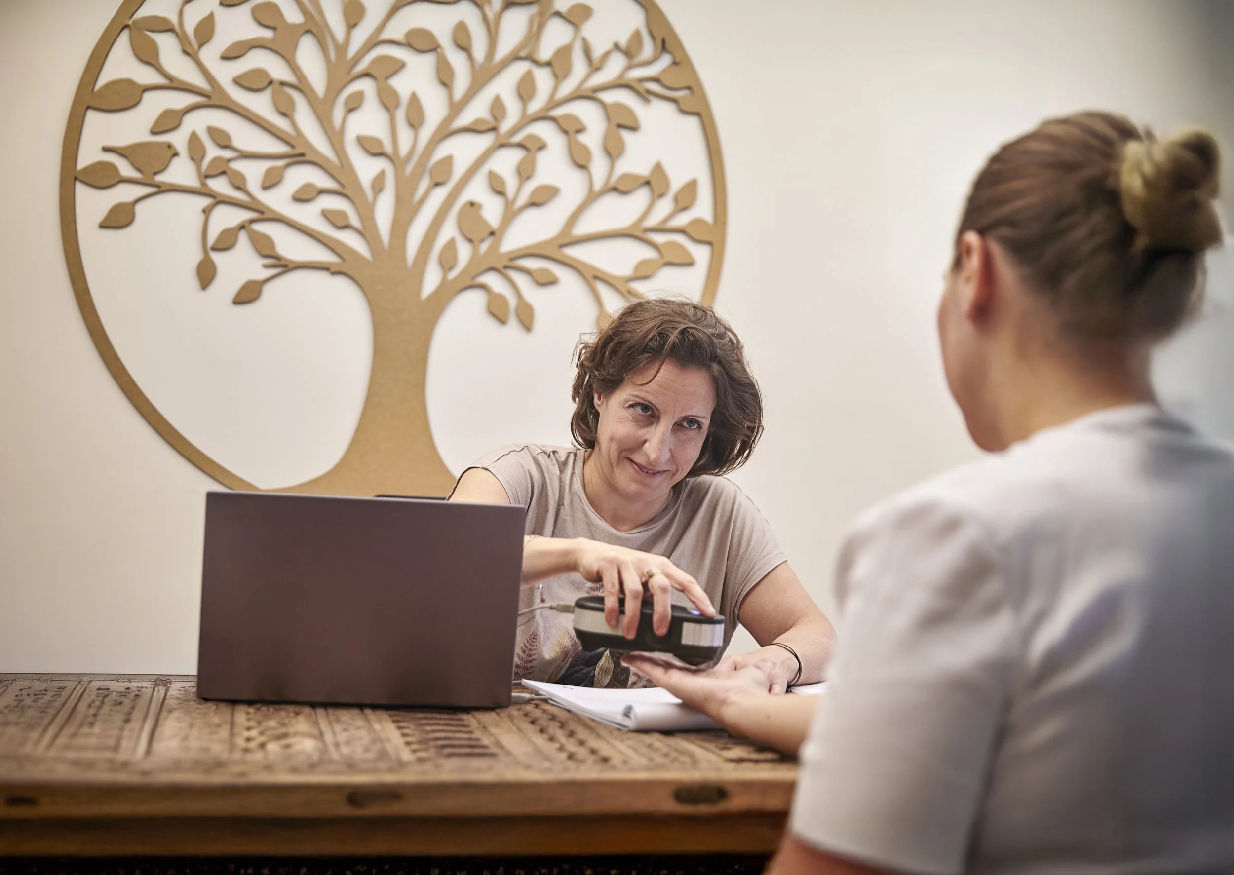 A woman handing over a payment device to another woman in a consultation or payment setting, with a laptop and documents on the table, and a decorative tree wall art in the background.