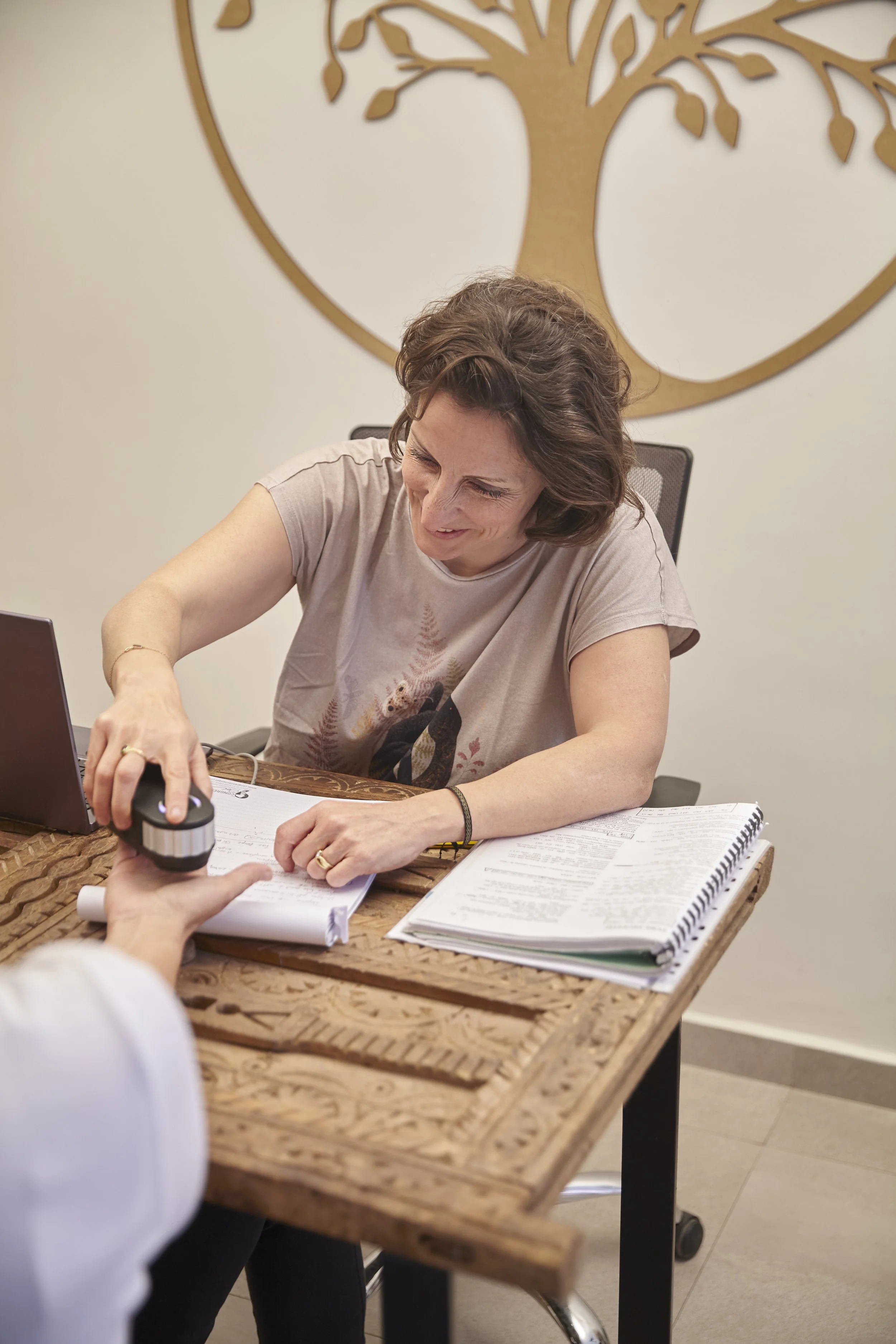 Woman smiling while being fingerprinted at a desk with papers, notebooks, and a laptop, in an office with a wall art of a tree.