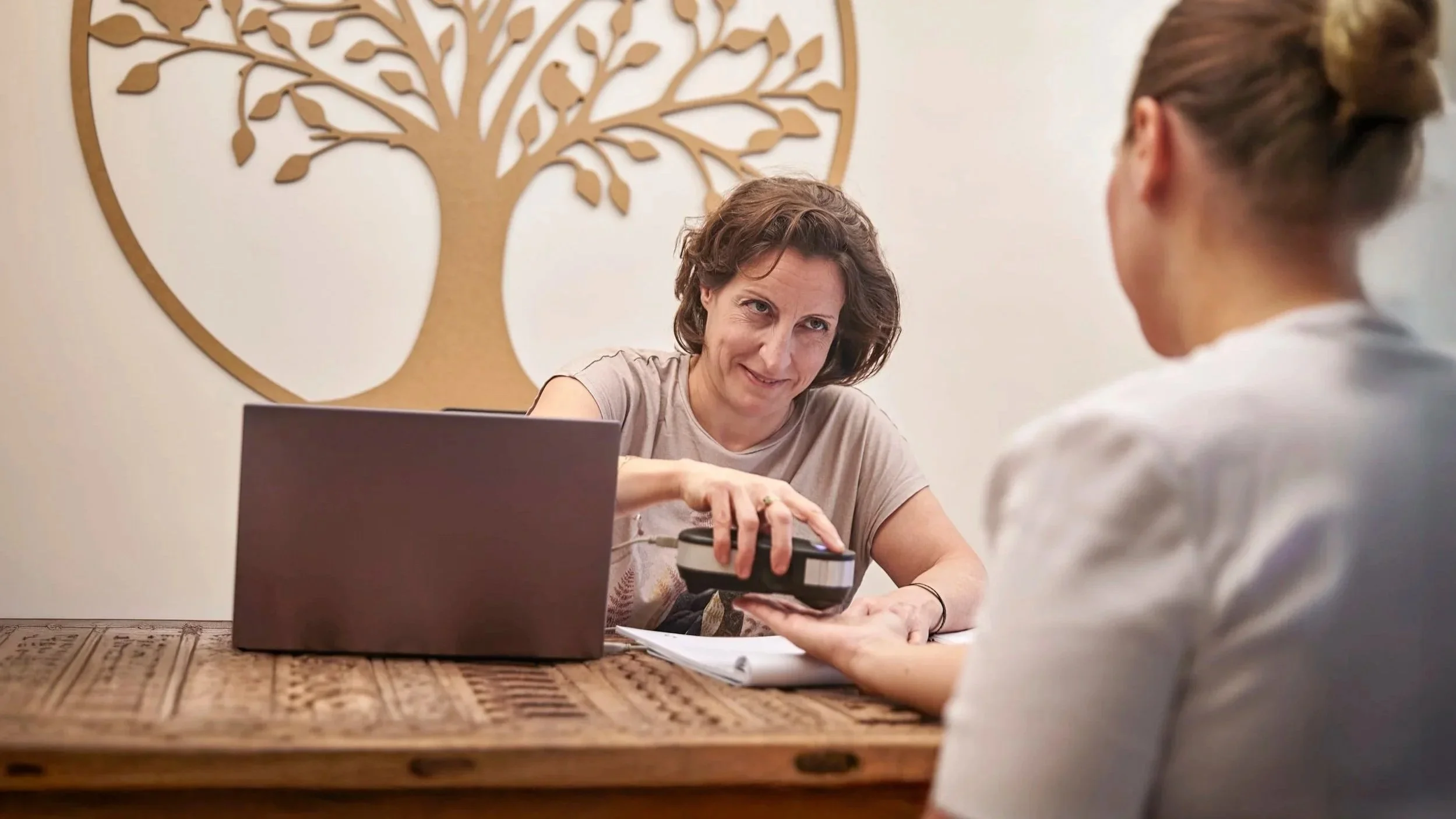 A woman at a desk handing an item to another woman, with a laptop, papers, and a wooden table, featuring a large wall art of a tree.