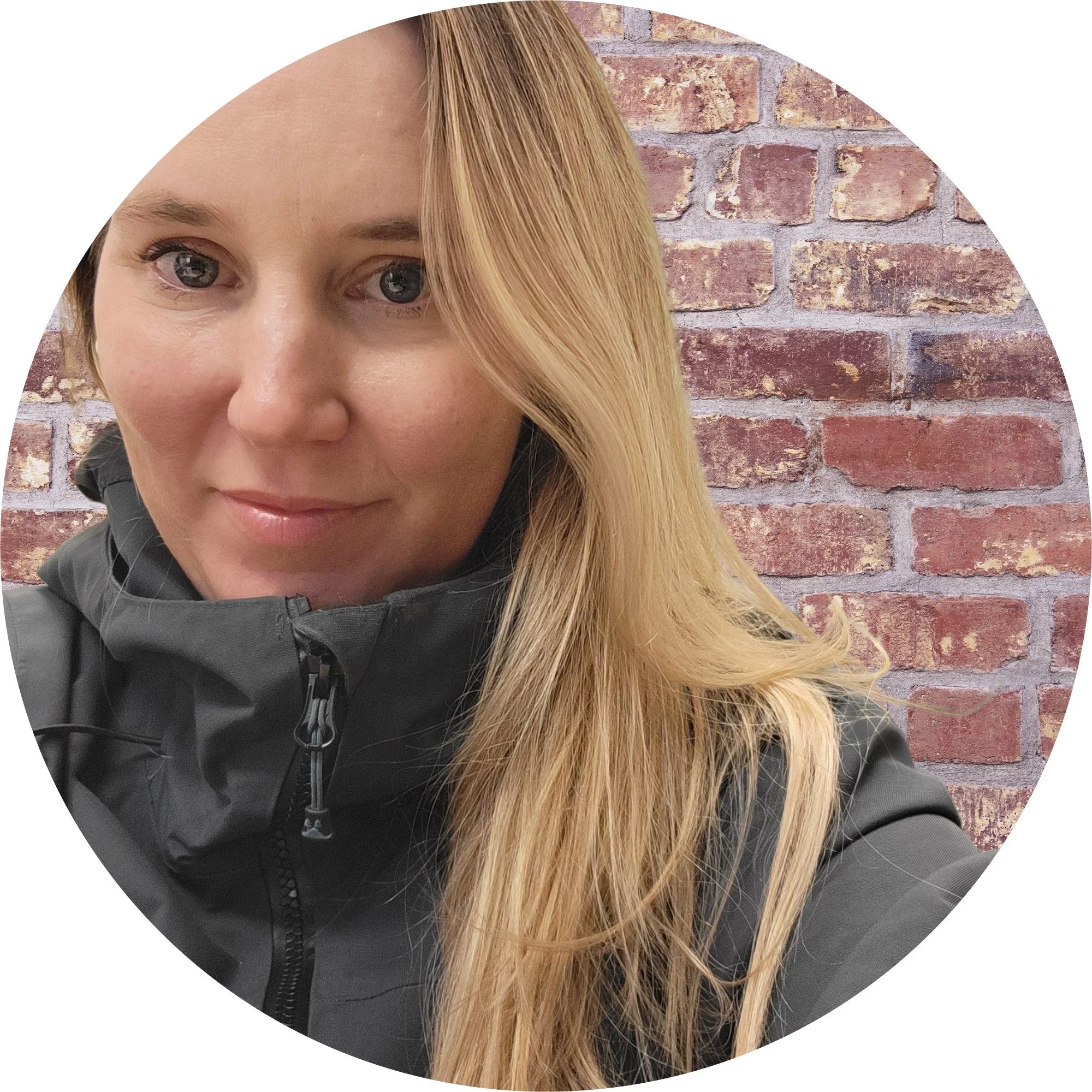 A woman with long blonde hair wearing a black jacket standing in front of a brick wall.