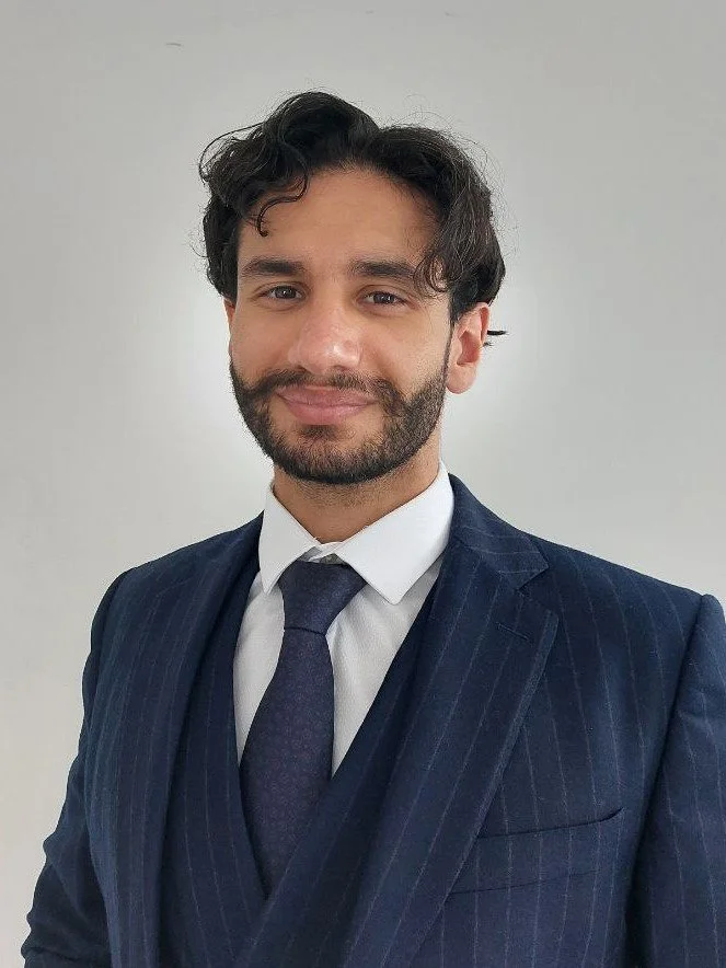 A young man with curly hair and a beard, dressed in a pinstripe suit, white shirt, and tie, smiling at the camera against a plain background.