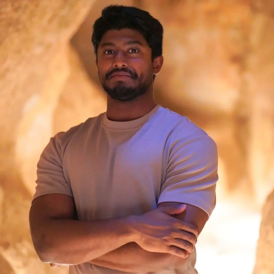 A man with dark hair, beard, and mustache standing with arms crossed in a rocky cave with warm lighting.