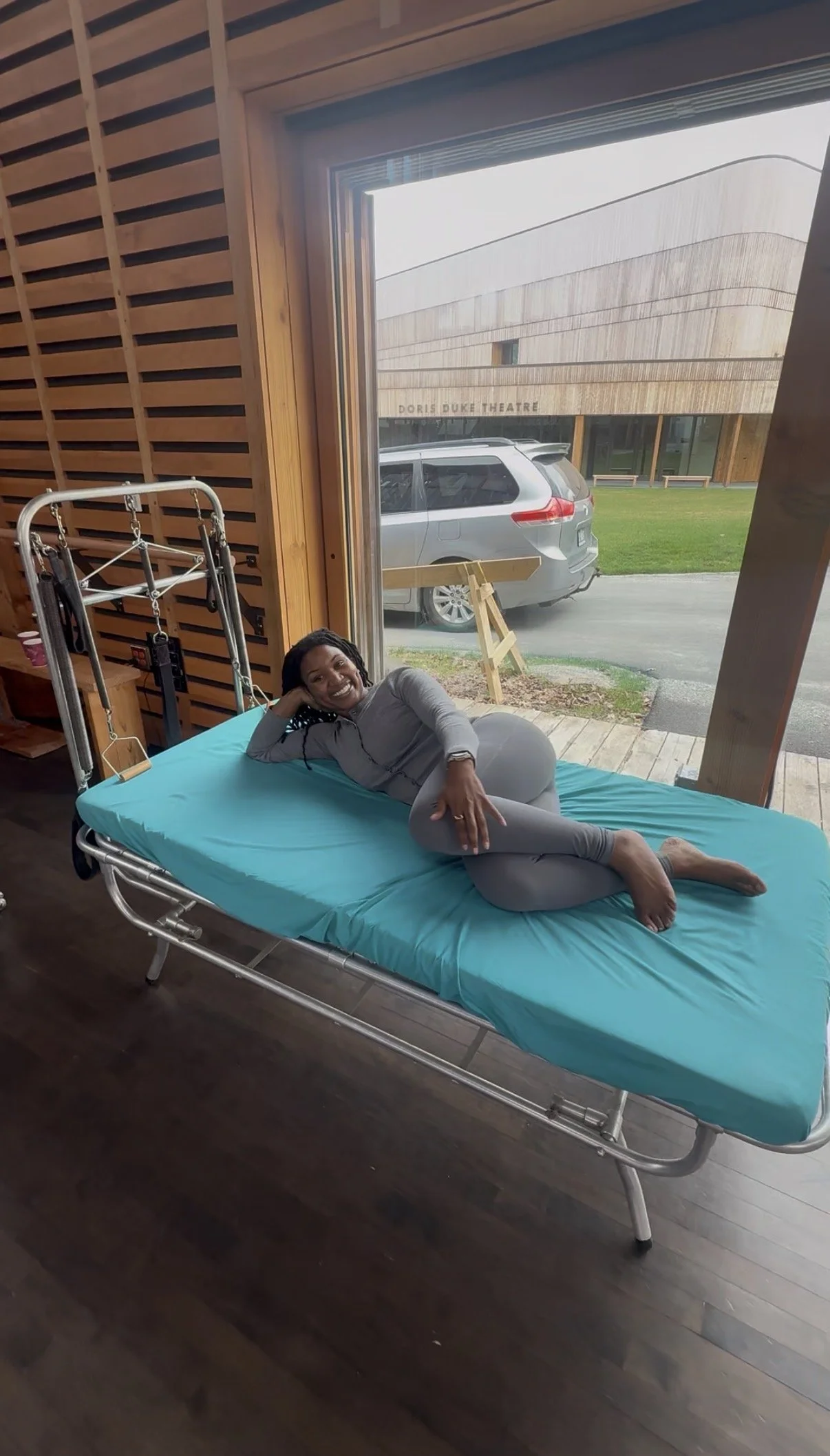 A woman lying on a hospital bed inside a wooden building, with a large open window showing a parking lot and a modern wooden building labeled 'Dore Duke Theatre.'